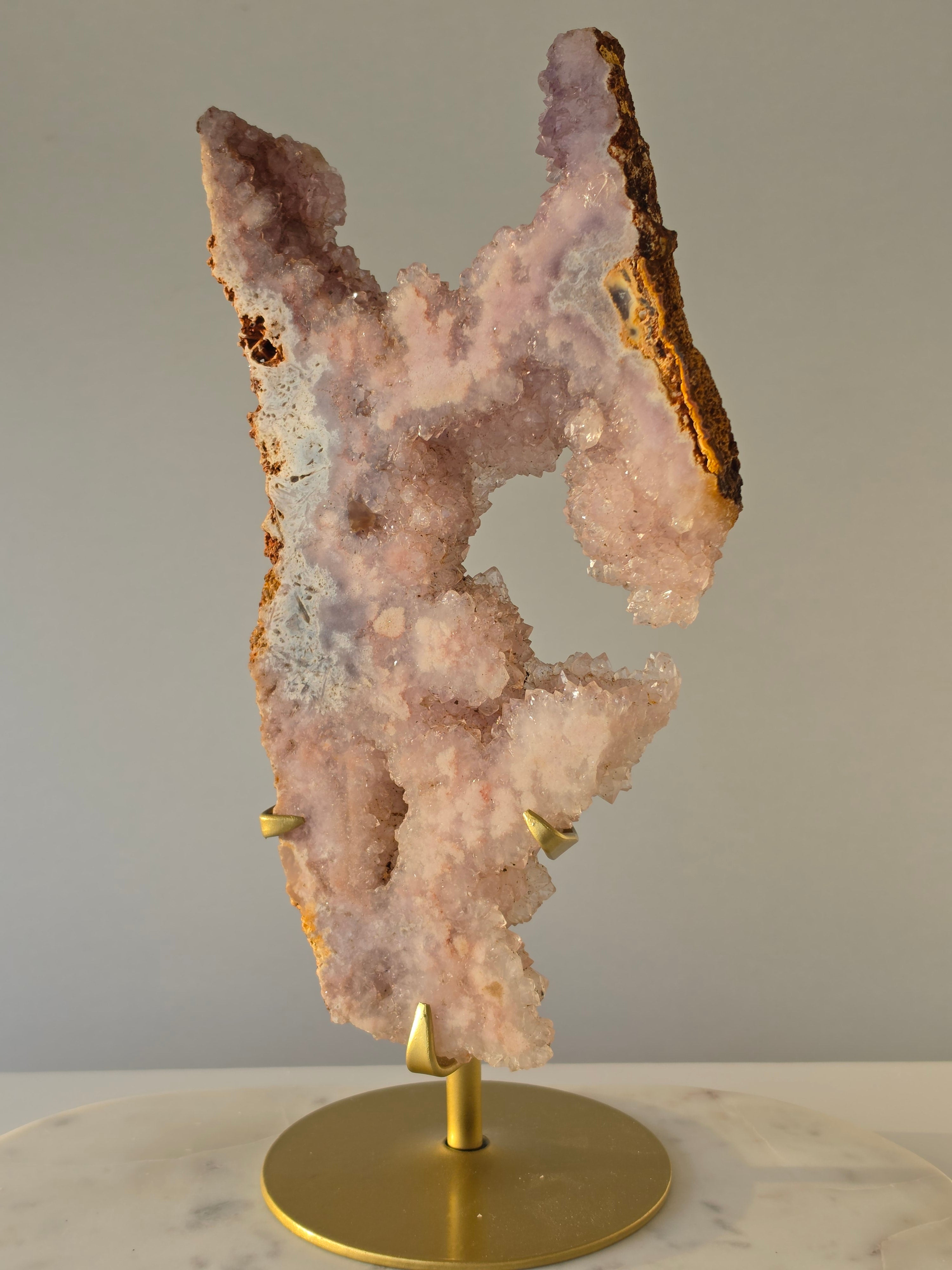 High-grade Brazilian Pink Amethyst slice on a gold display stand featuring sparkling blush-pink druzy crystals, natural agate banding, sculptural open formations, and delicate rose and champagne tones, photographed in natural light