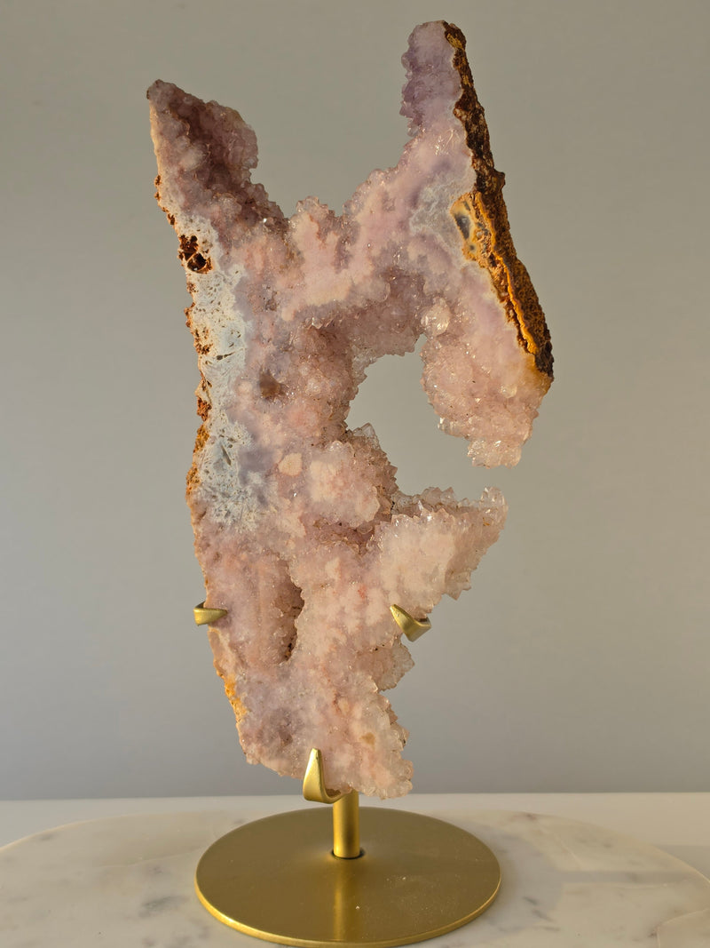 High-grade Brazilian Pink Amethyst slice on a gold display stand featuring sparkling blush-pink druzy crystals, natural agate banding, sculptural open formations, and delicate rose and champagne tones, photographed in natural light