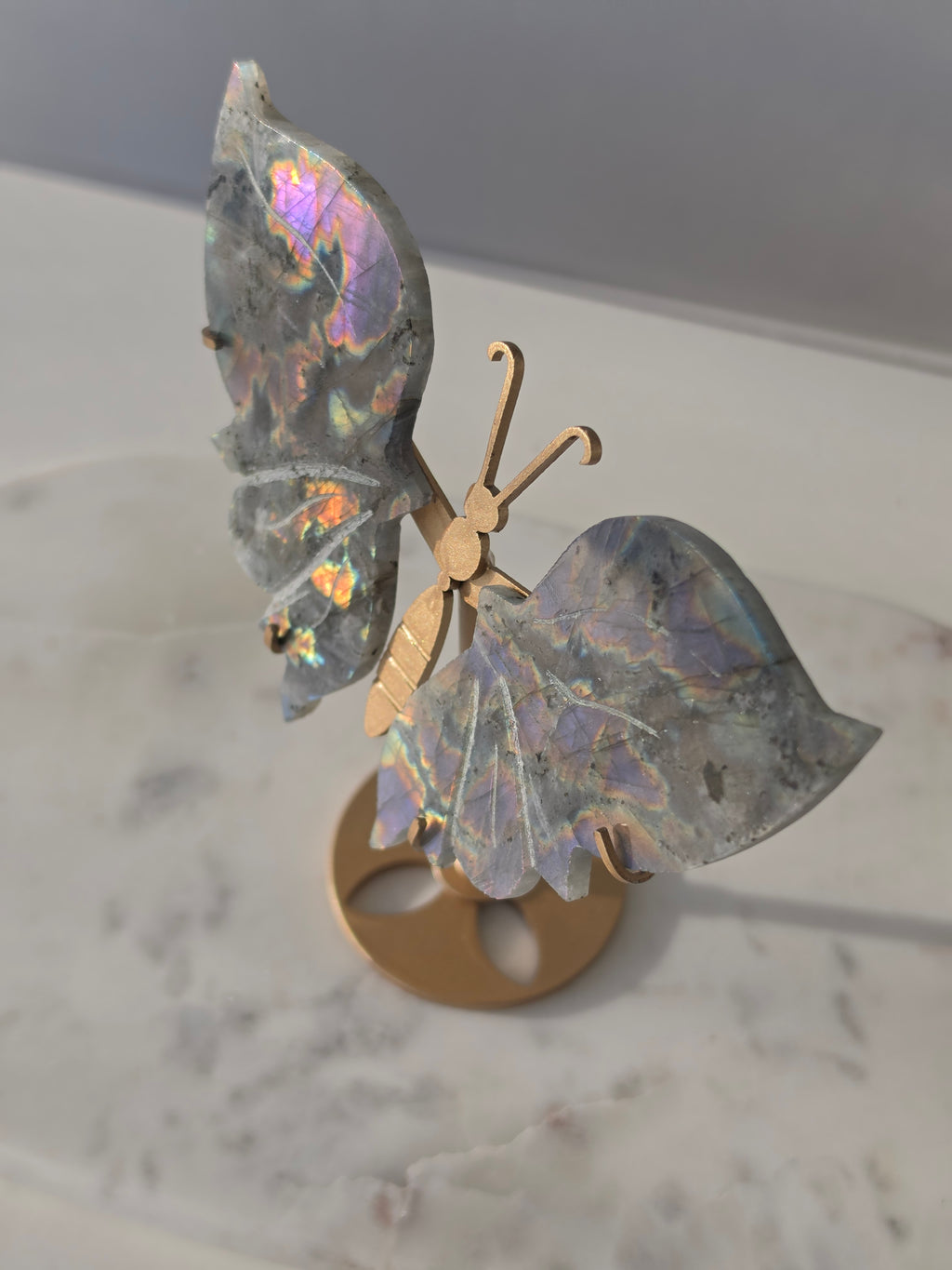 Labradorite butterfly crystal carving on a gold-toned stand with vibrant purple, blue, aqua and orange iridescent rainbow flashes across its grey stone wings.