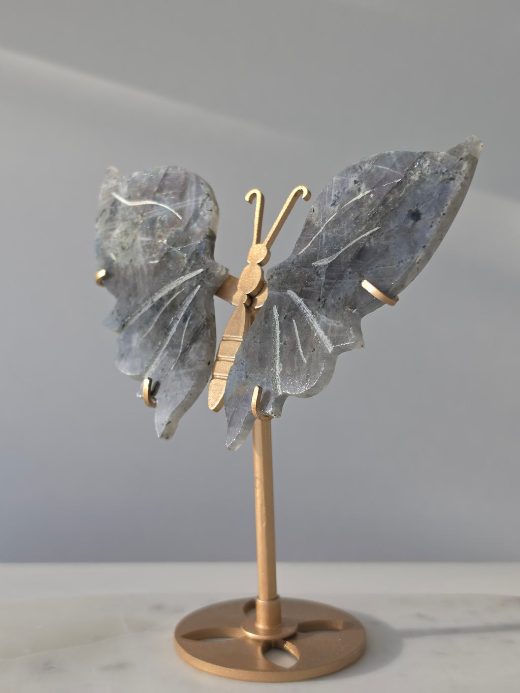 Labradorite butterfly crystal carving on a gold-toned stand with vibrant purple, blue, aqua and orange iridescent rainbow flashes across its grey stone wings.