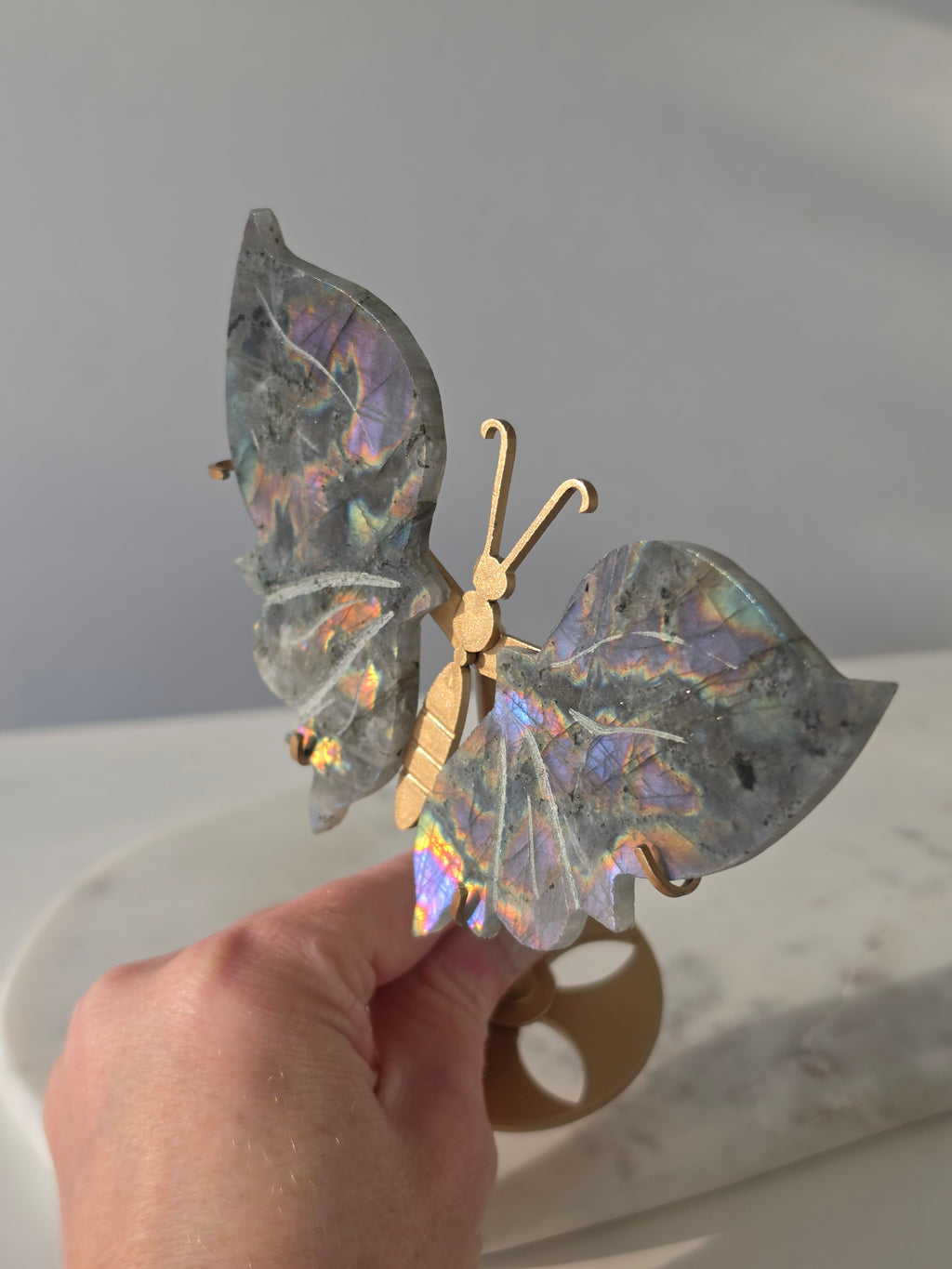 Labradorite butterfly crystal carving on a gold-toned stand with vibrant purple, blue, aqua and orange iridescent rainbow flashes across its grey stone wings.