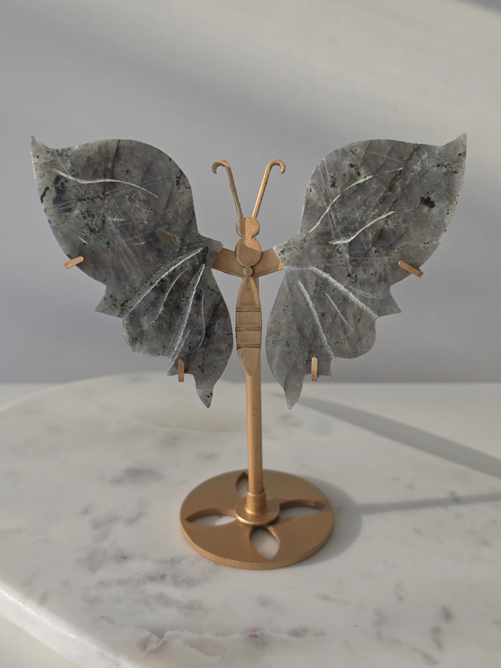 Labradorite butterfly crystal carving on a gold-toned stand with vibrant purple, blue, aqua and orange iridescent rainbow flashes across its grey stone wings.