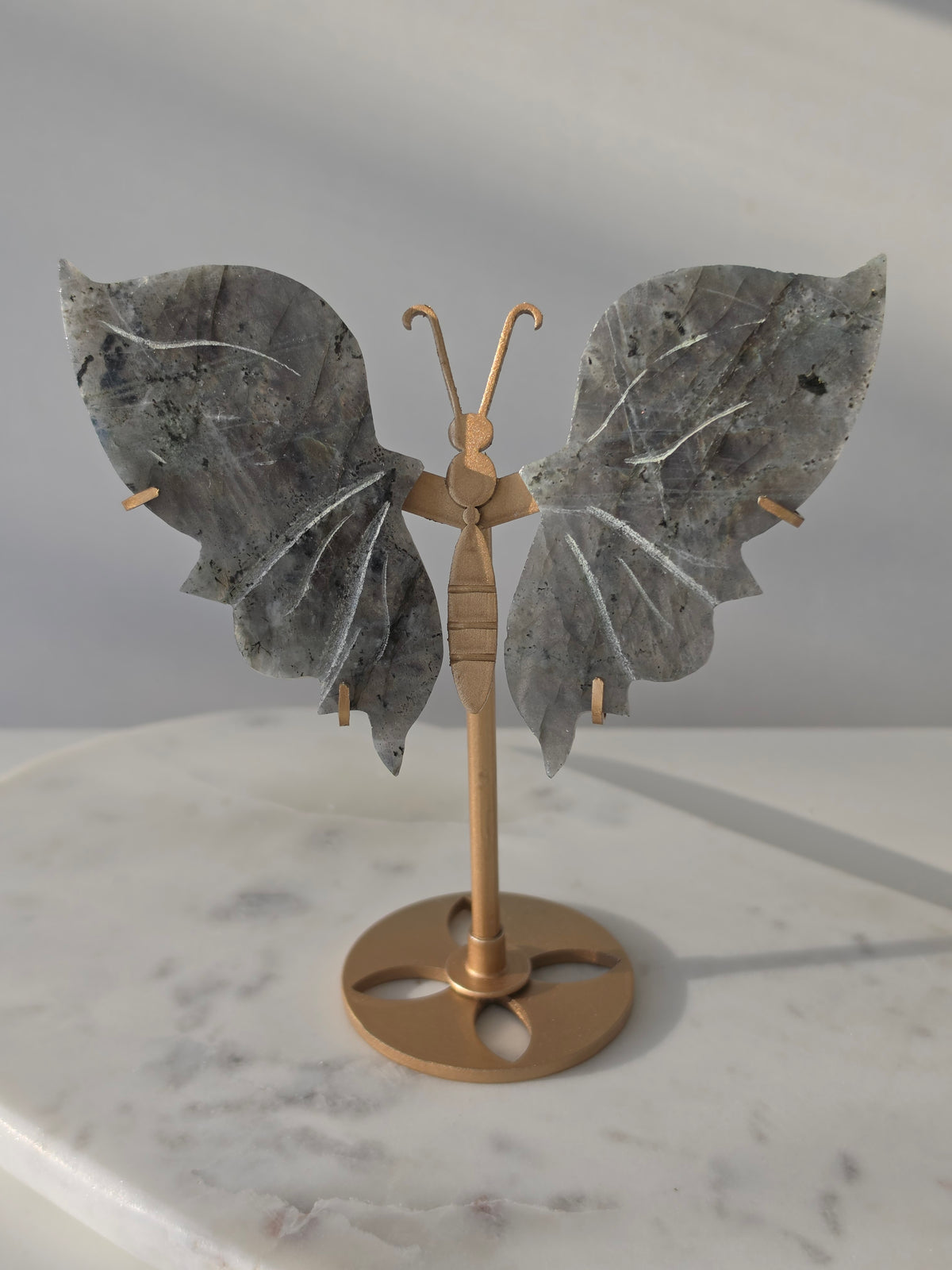 Labradorite butterfly crystal carving on a gold-toned stand with vibrant purple, blue, aqua and orange iridescent rainbow flashes across its grey stone wings.