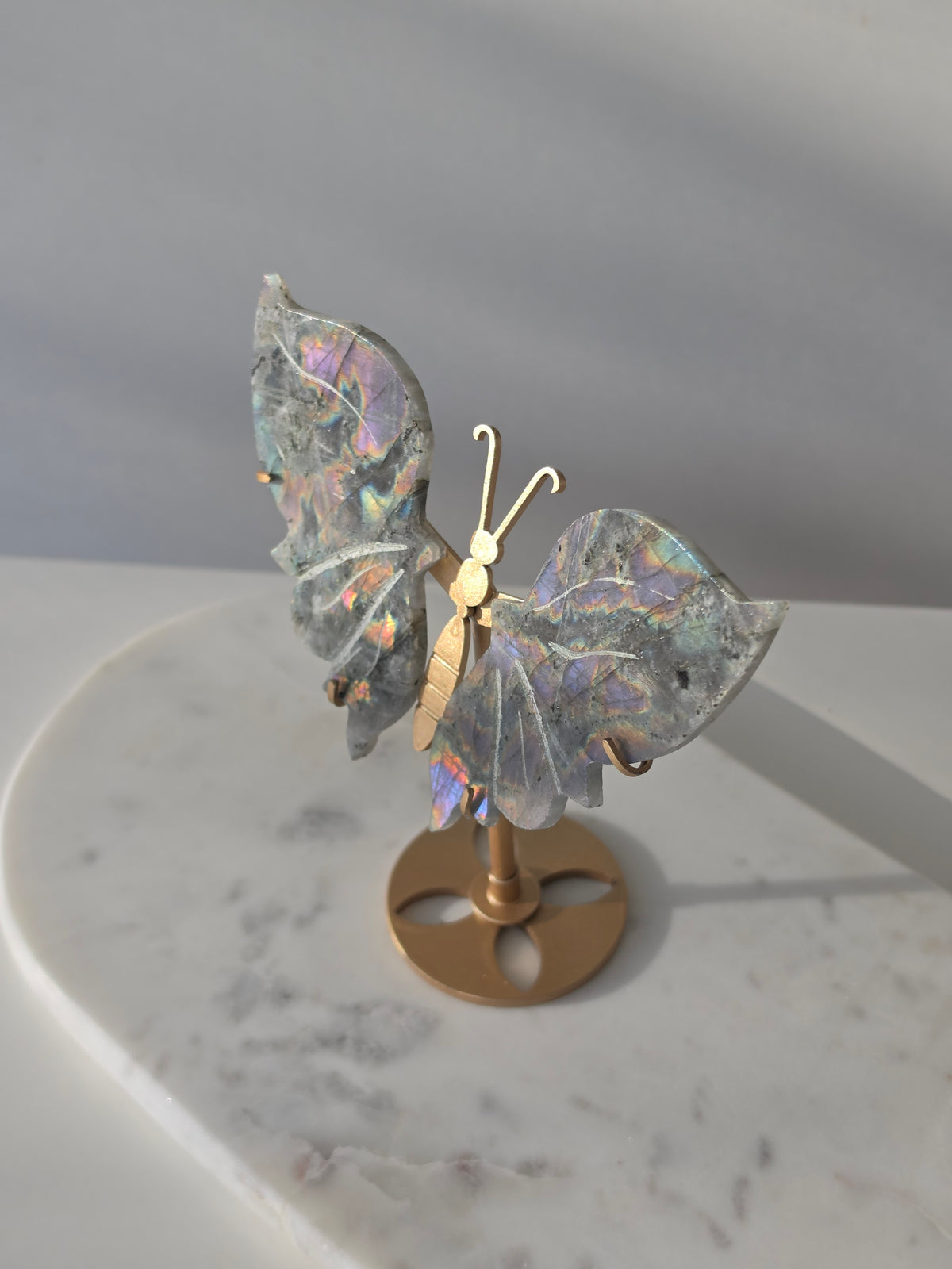 Labradorite butterfly crystal carving on a gold-toned stand with vibrant purple, blue, aqua and orange iridescent rainbow flashes across its grey stone wings.