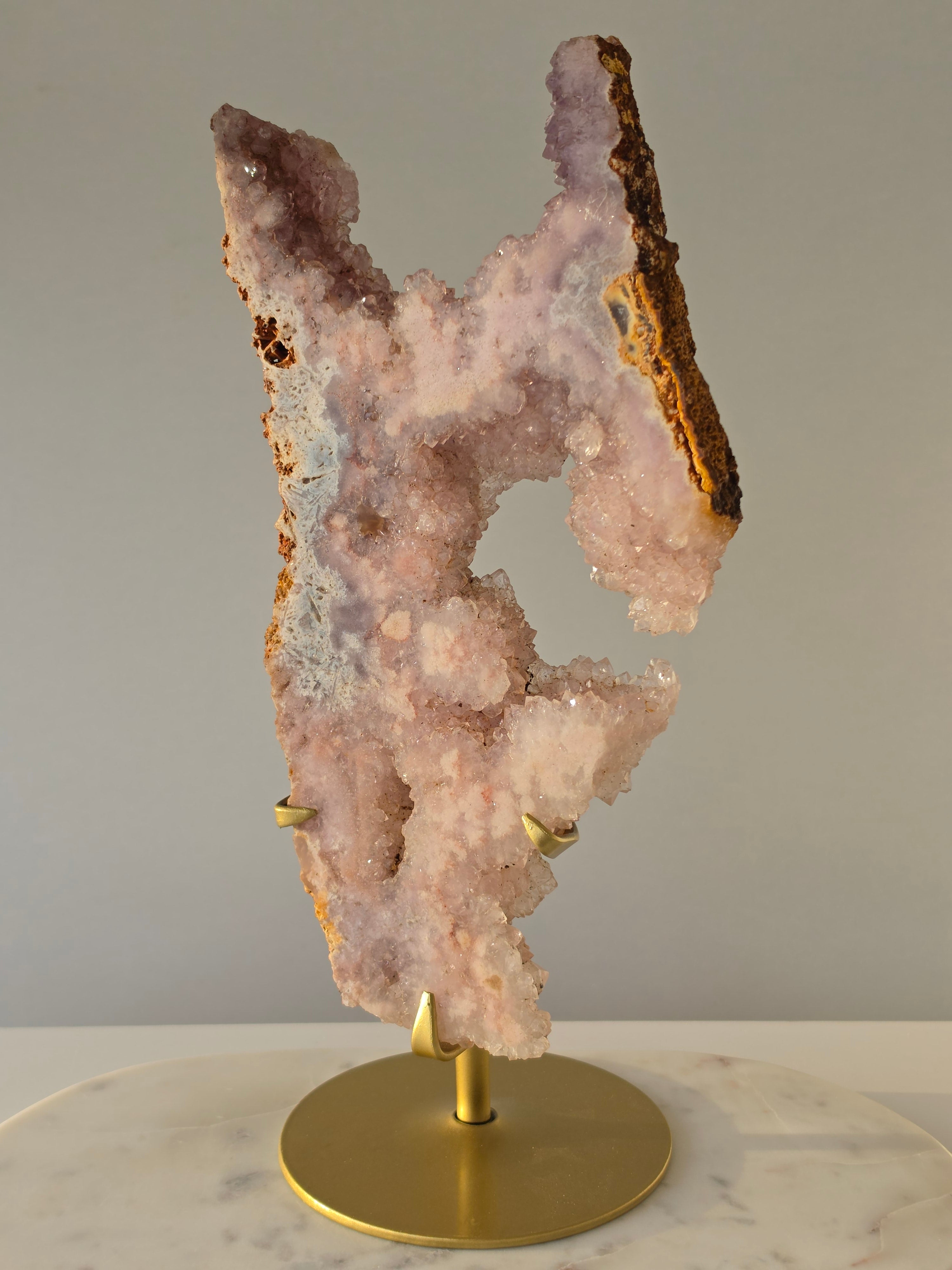 High-grade Brazilian Pink Amethyst slice on a gold display stand featuring sparkling blush-pink druzy crystals, natural agate banding, sculptural open formations, and delicate rose and champagne tones, photographed in natural light