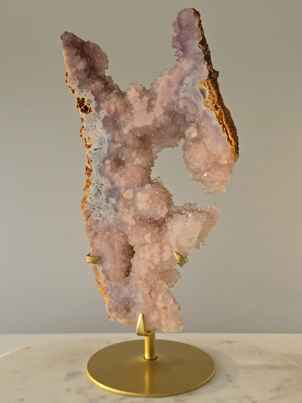 High-grade Brazilian Pink Amethyst slice on a gold display stand featuring sparkling blush-pink druzy crystals, natural agate banding, sculptural open formations, and delicate rose and champagne tones, photographed in natural light