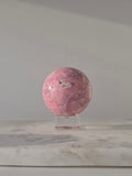 High-grade Peruvian Pink Rhodonite crystal sphere featuring vibrant raspberry and blush-pink tones, soft cream marbling, natural black manganese veining, and a flawless high-polish finish, displayed on a clear acrylic stand in natural light.