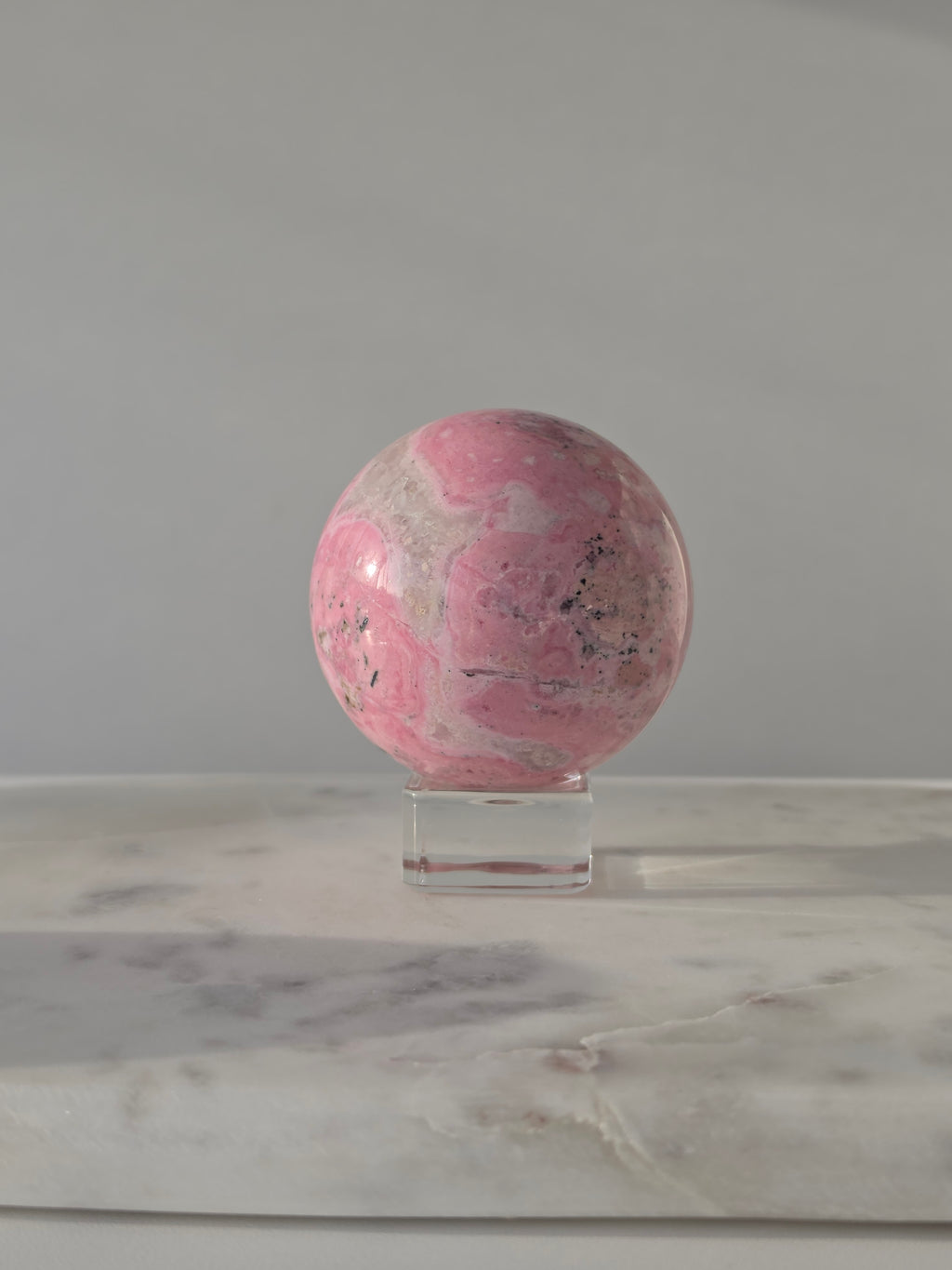 High-grade Peruvian Pink Rhodonite crystal sphere featuring vibrant raspberry and blush-pink tones, soft cream marbling, natural black manganese veining, and a flawless high-polish finish, displayed on a clear acrylic stand in natural light.
