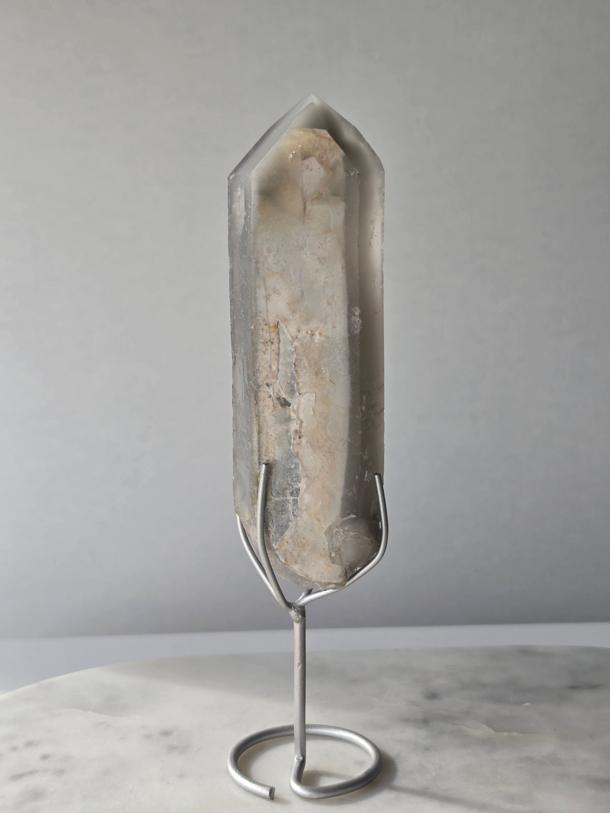 Translucent grey lithium crystal point with delicate internal phantom-like inclusions displayed on a silver-toned metal stand, 337g and approximately 20cm high.