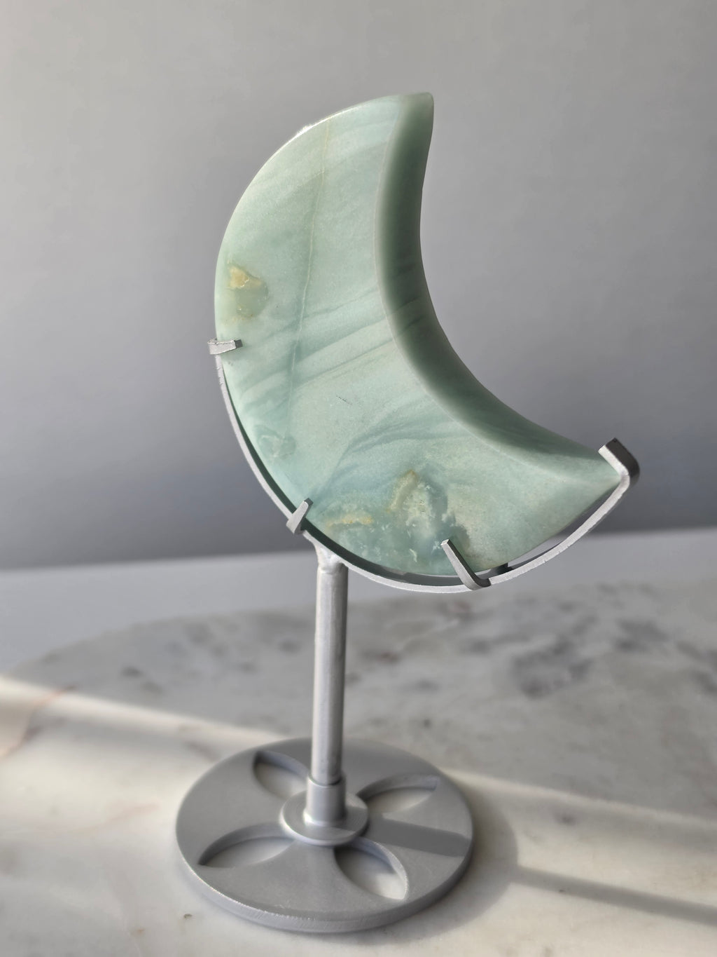 Natural Amazonite crescent moon with soft seafoam green and blue-green banding, subtle golden-brown inclusions and polished finish, displayed on a silver stand.