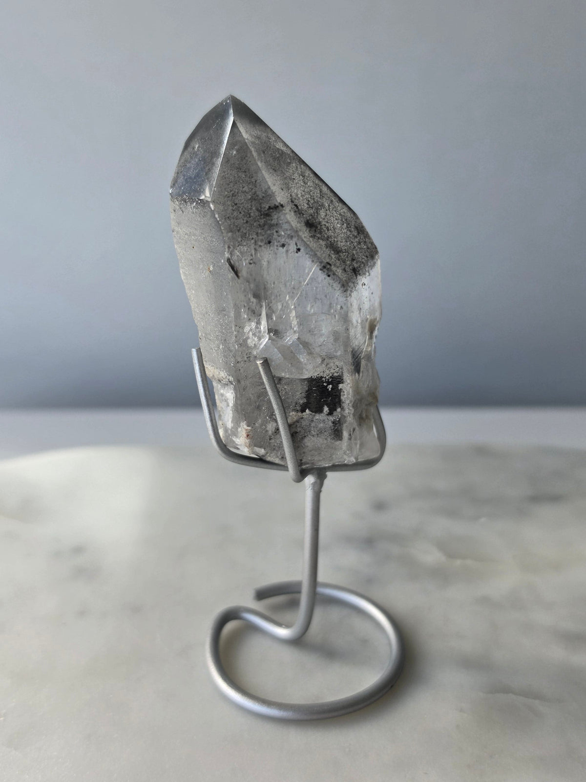 Black Phantom Lemurian Seed quartz point featuring smoky translucent crystal and dramatic charcoal-black internal phantom-like formations, displayed on a silver-toned stand.