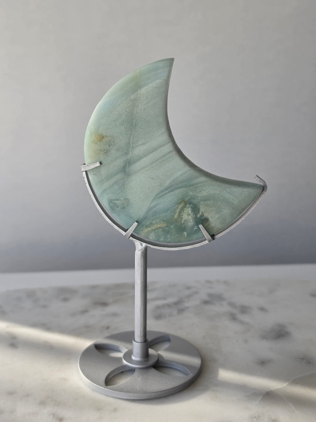 Natural Amazonite crescent moon with soft seafoam green and blue-green banding, subtle golden-brown inclusions and polished finish, displayed on a silver stand.