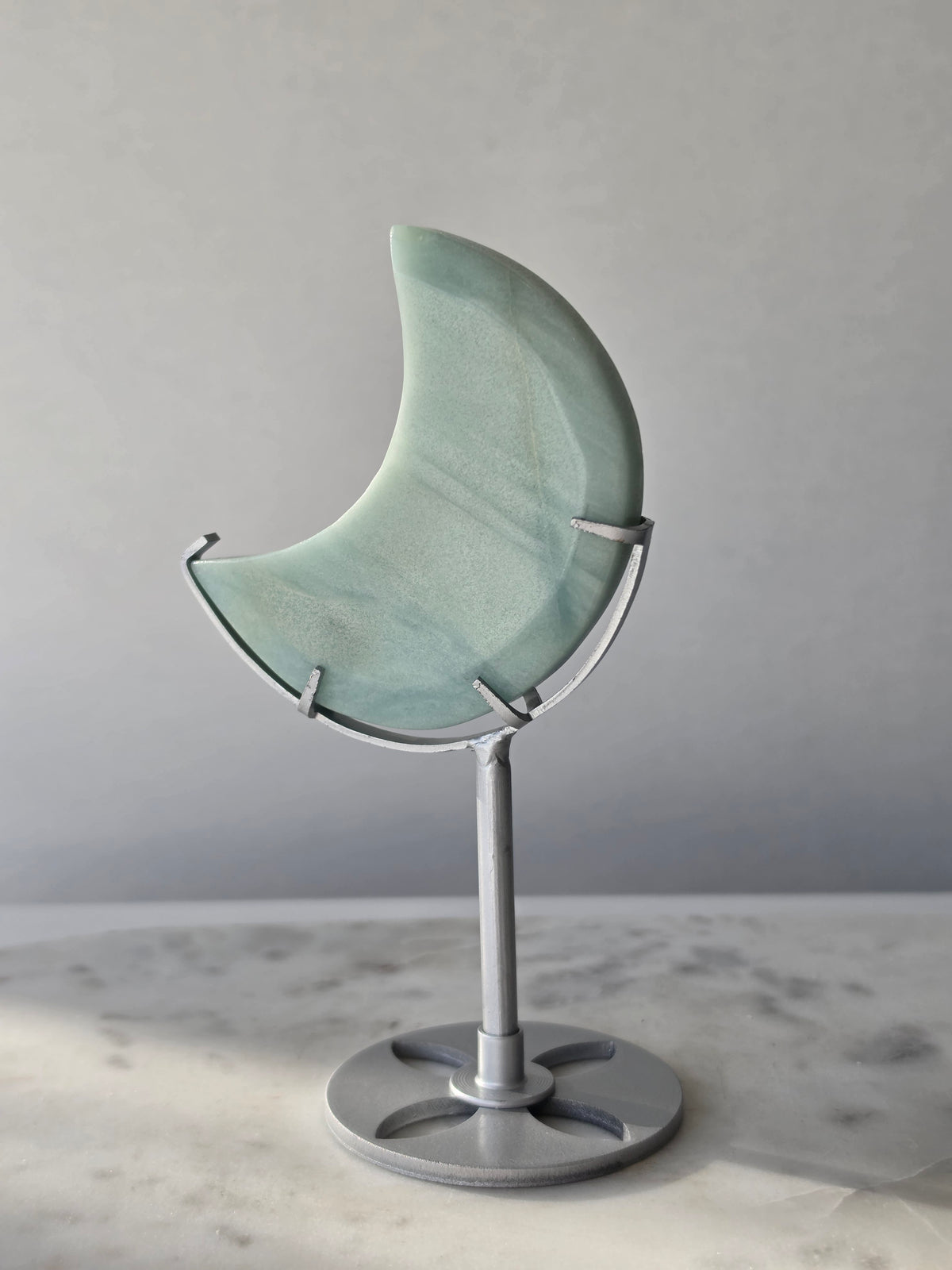 Natural Amazonite crescent moon with soft seafoam green and blue-green banding, subtle golden-brown inclusions and polished finish, displayed on a silver stand.