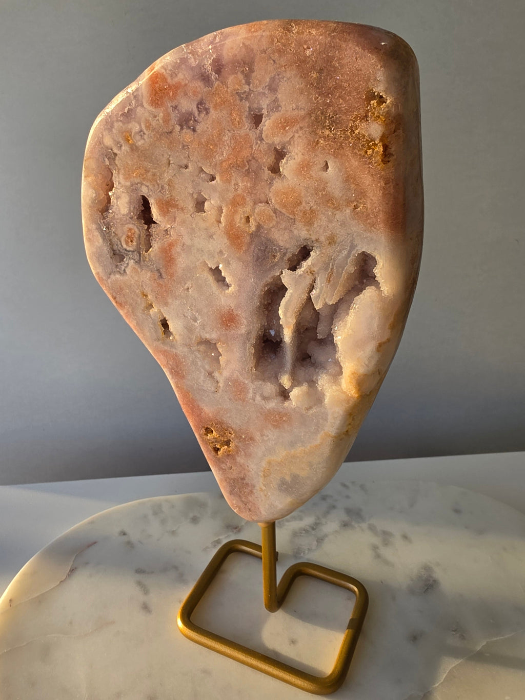 Pink Amethyst statement slab with soft blush pink and lavender tones, unique druzy bubble formations and sparkling crystal pockets, displayed on an elegant gold-toned stand.
