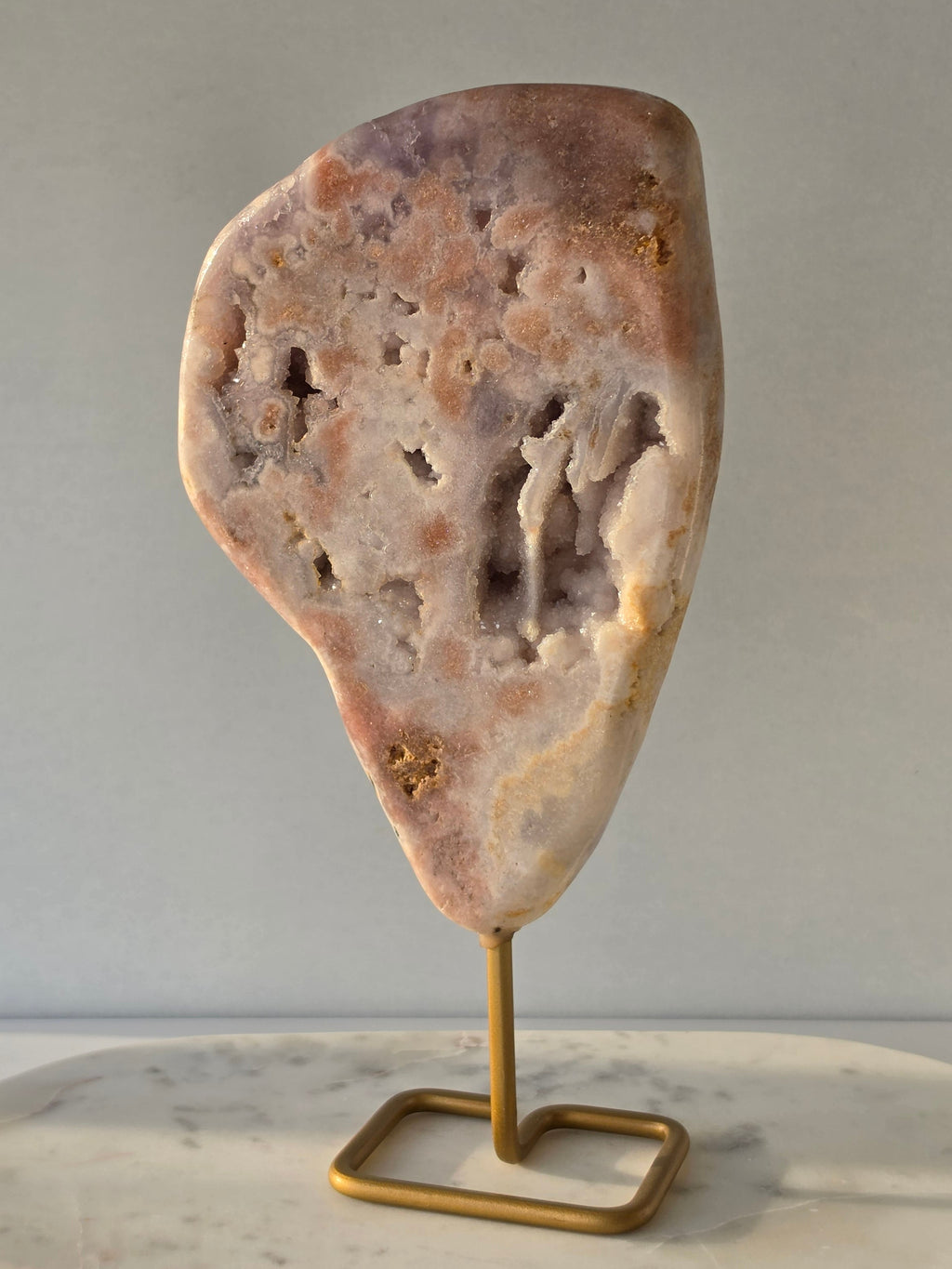 Pink Amethyst statement slab with soft blush pink and lavender tones, unique druzy bubble formations and sparkling crystal pockets, displayed on an elegant gold-toned stand.