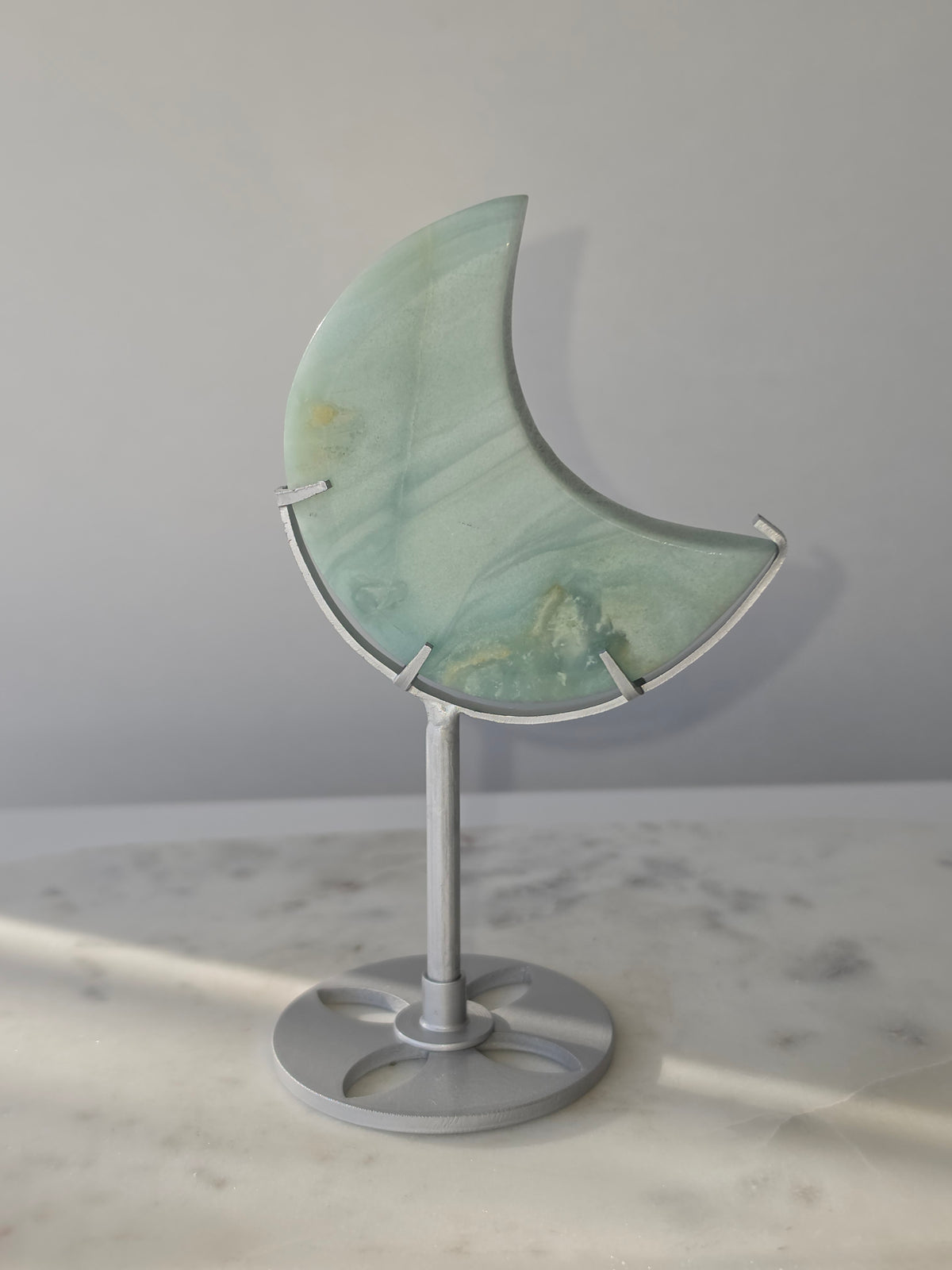 Natural Amazonite crescent moon with soft seafoam green and blue-green banding, subtle golden-brown inclusions and polished finish, displayed on a silver stand.