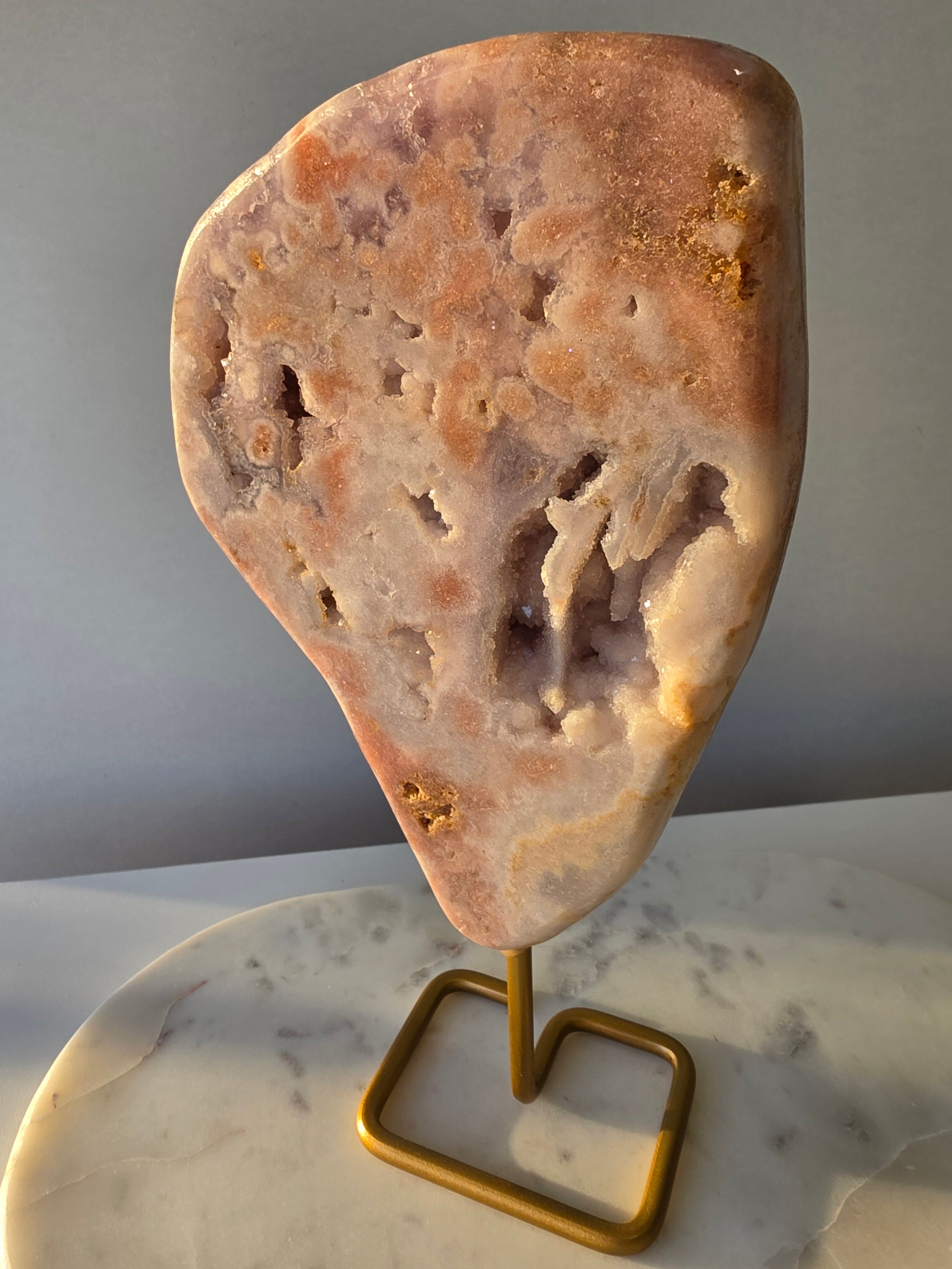 Pink Amethyst statement slab with soft blush pink and lavender tones, unique druzy bubble formations and sparkling crystal pockets, displayed on an elegant gold-toned stand.
