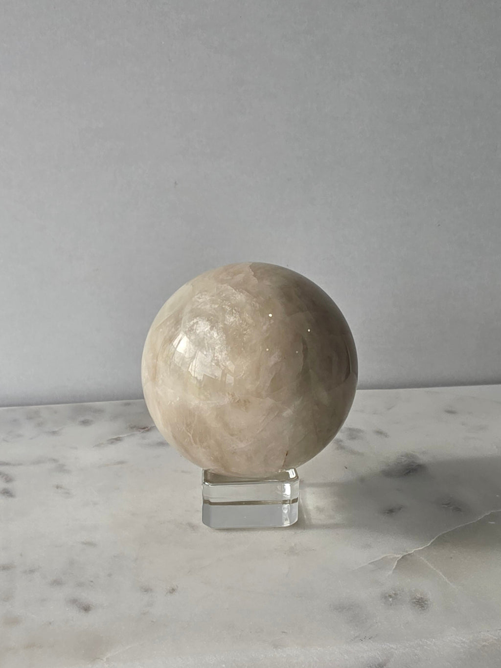 Polished Moonstone Sphere in creamy ivory, champagne and taupe-grey tones, displaying subtle natural patterns and a soft luminous sheen on a clear stand.