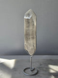Translucent grey lithium crystal point with delicate internal phantom-like inclusions displayed on a silver-toned metal stand, 337g and approximately 20cm high.