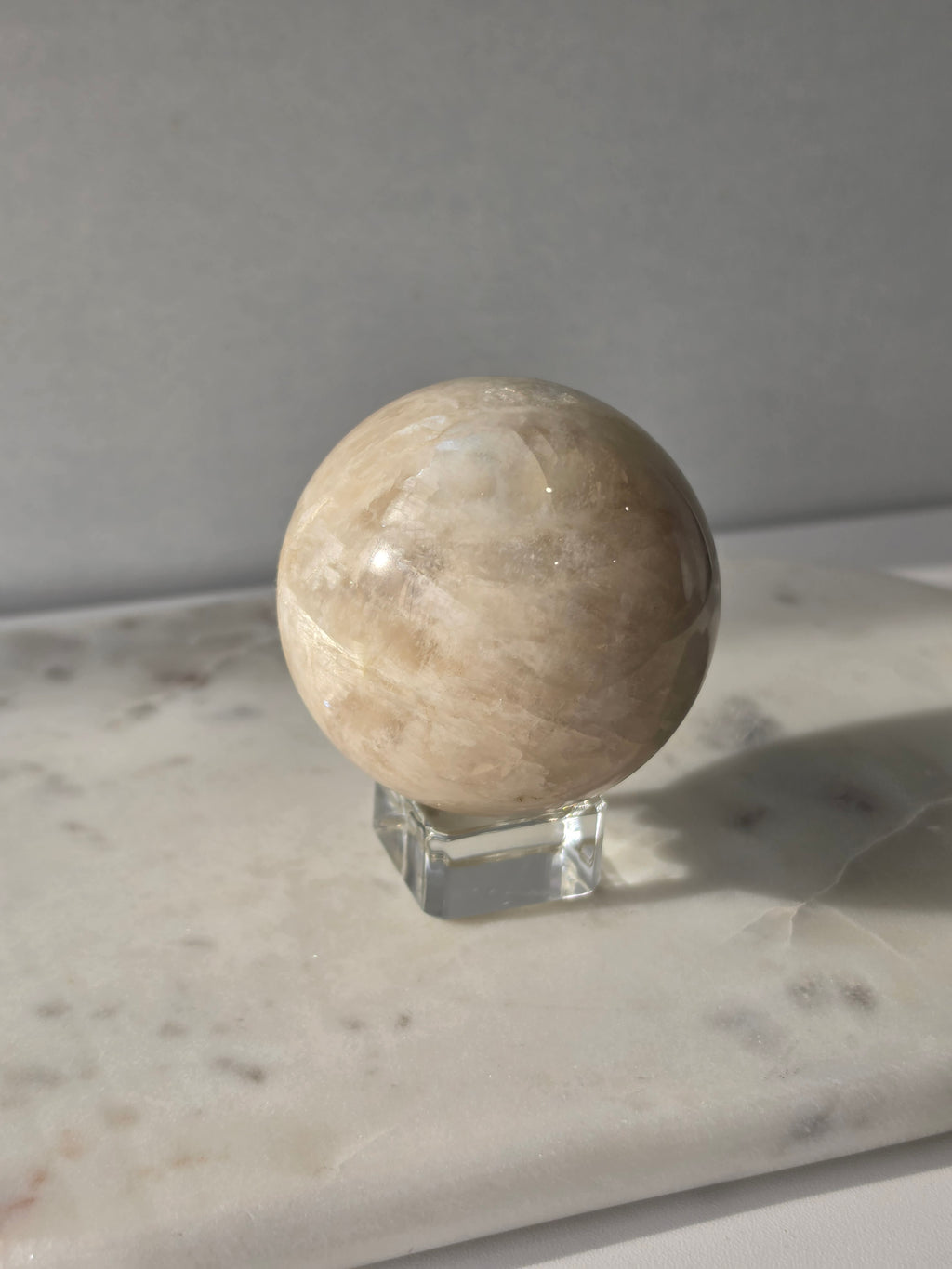 Polished Moonstone Sphere in creamy ivory, champagne and taupe-grey tones, displaying subtle natural patterns and a soft luminous sheen on a clear stand.