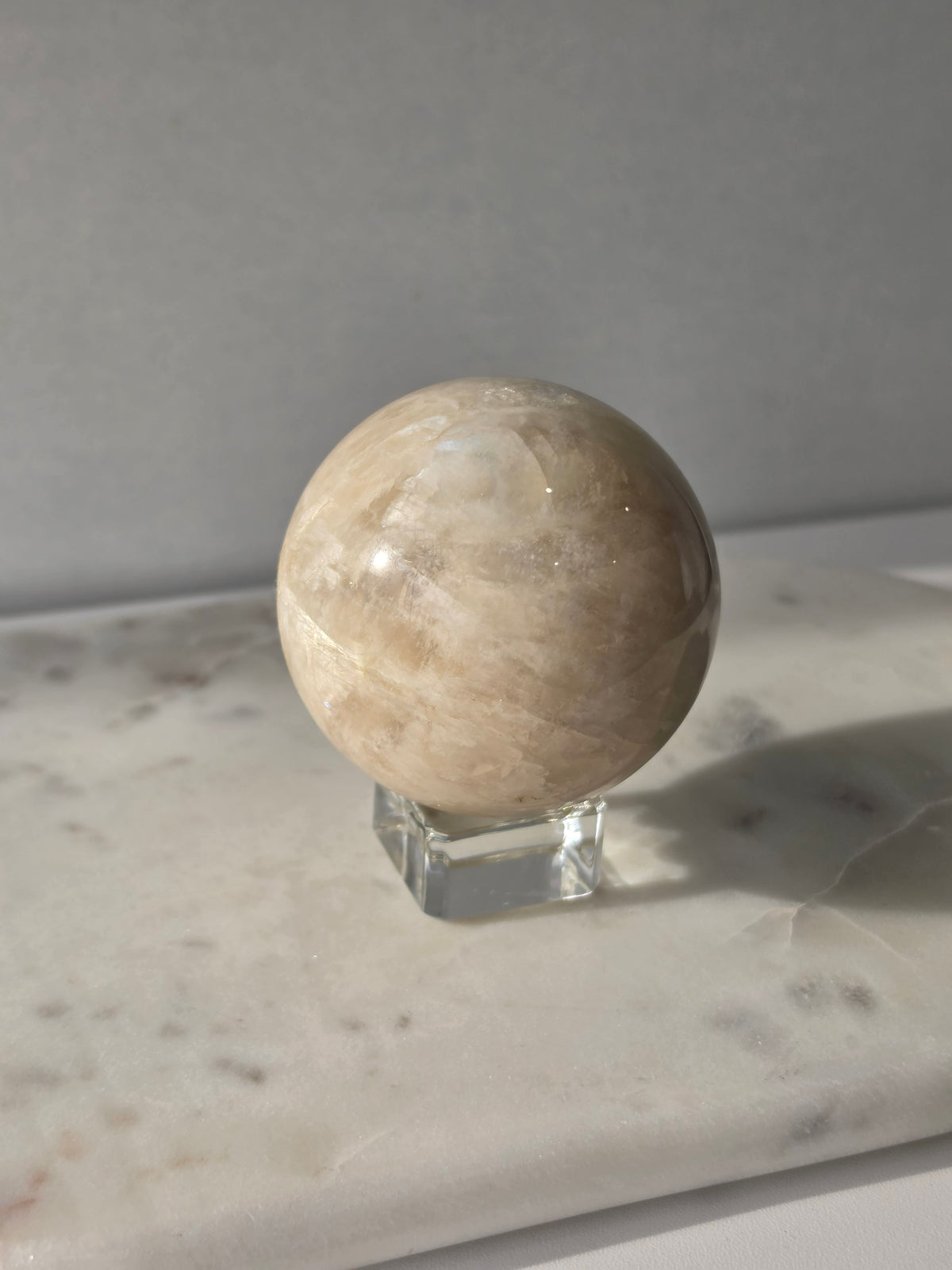Polished Moonstone Sphere in creamy ivory, champagne and taupe-grey tones, displaying subtle natural patterns and a soft luminous sheen on a clear stand.