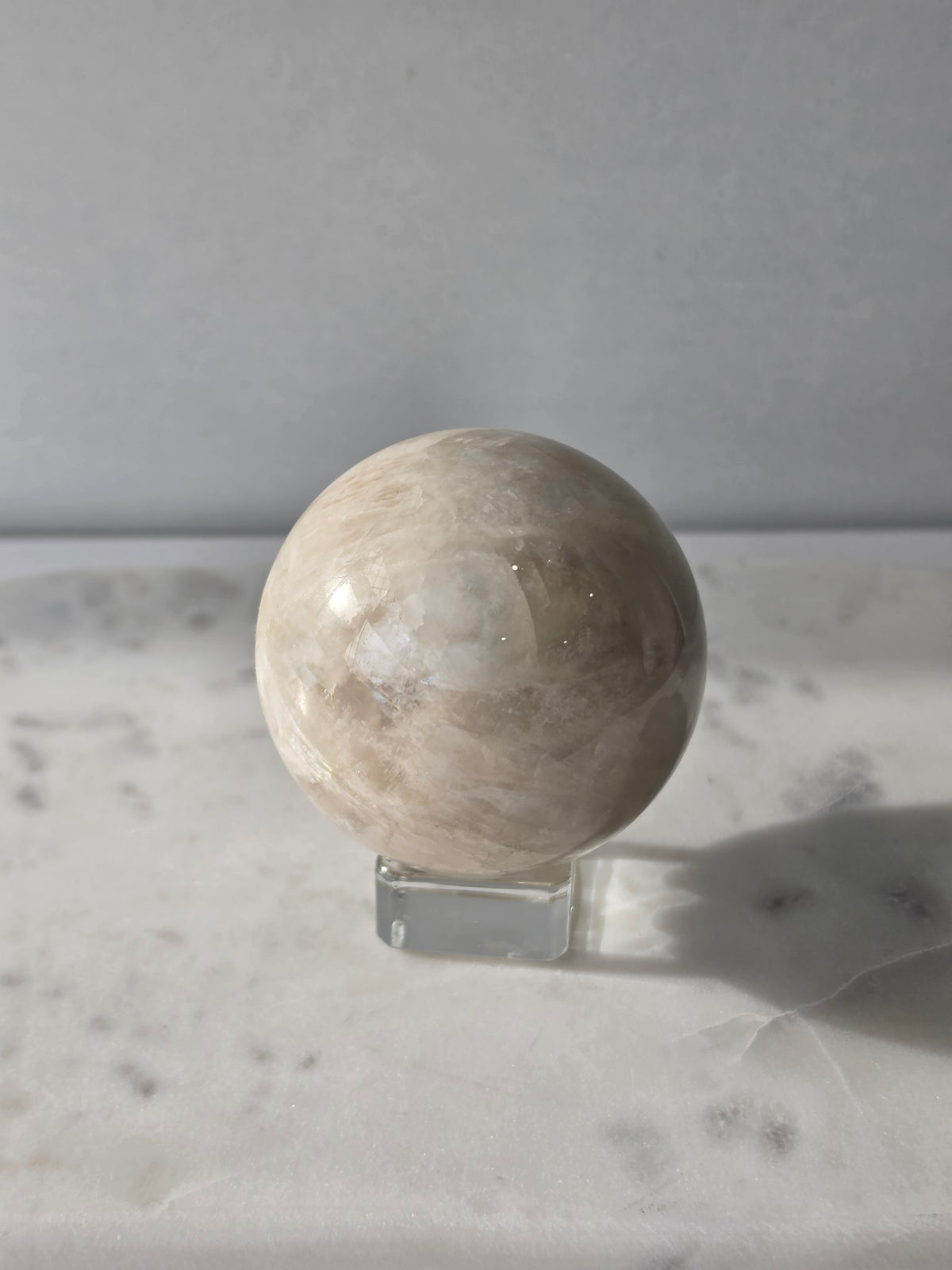Polished Moonstone Sphere in creamy ivory, champagne and taupe-grey tones, displaying subtle natural patterns and a soft luminous sheen on a clear stand.