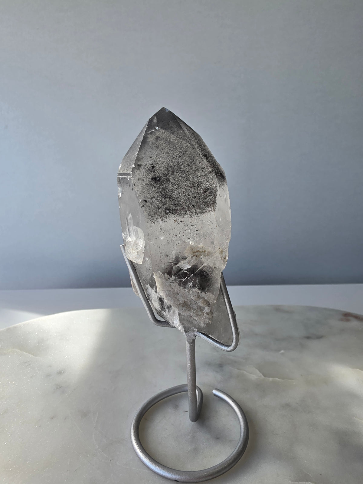 Black Phantom Lemurian Seed quartz point featuring smoky translucent crystal and dramatic charcoal-black internal phantom-like formations, displayed on a silver-toned stand.