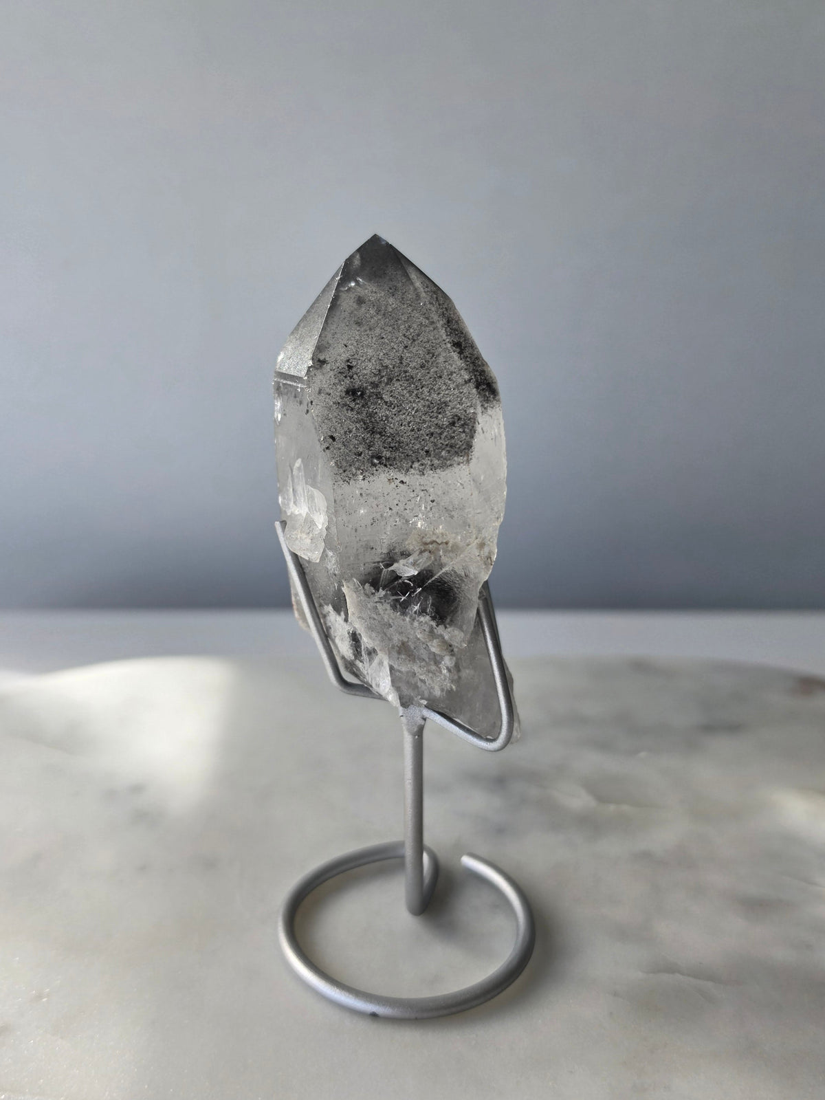 Black Phantom Lemurian Seed quartz point featuring smoky translucent crystal and dramatic charcoal-black internal phantom-like formations, displayed on a silver-toned stand.