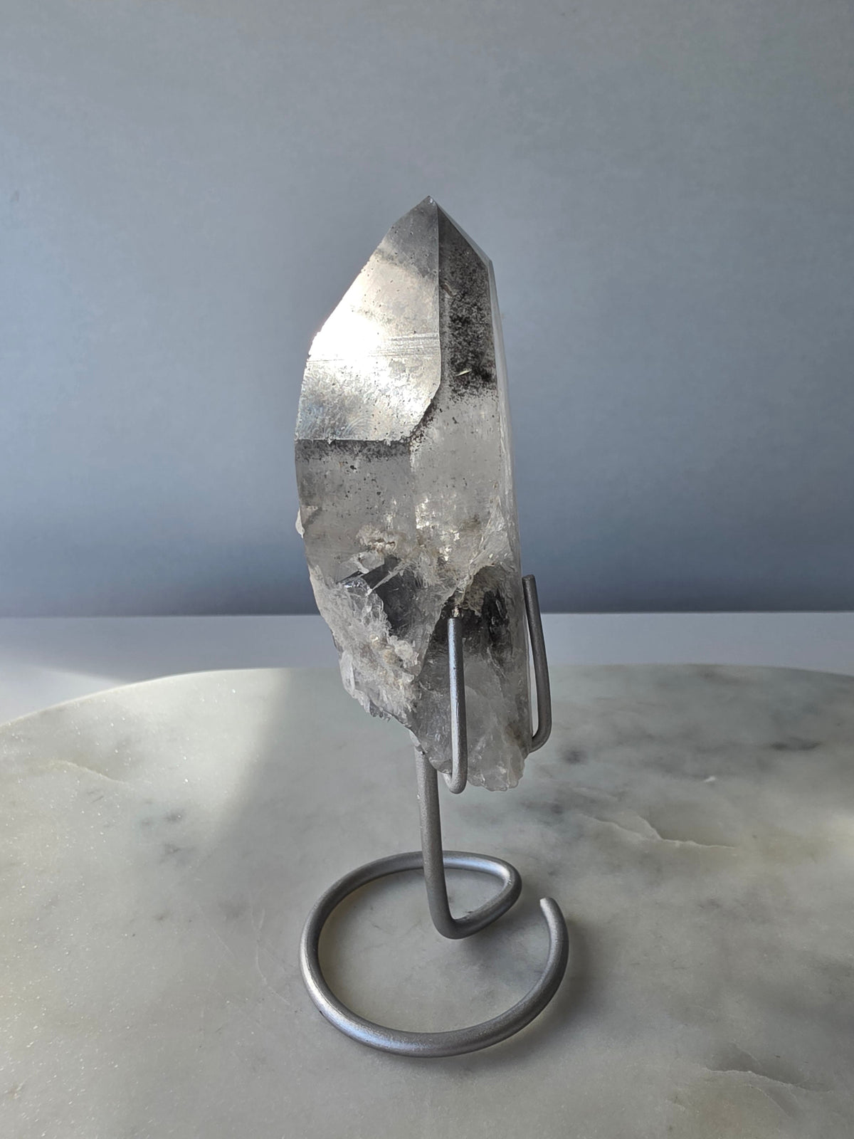 Black Phantom Lemurian Seed quartz point featuring smoky translucent crystal and dramatic charcoal-black internal phantom-like formations, displayed on a silver-toned stand.