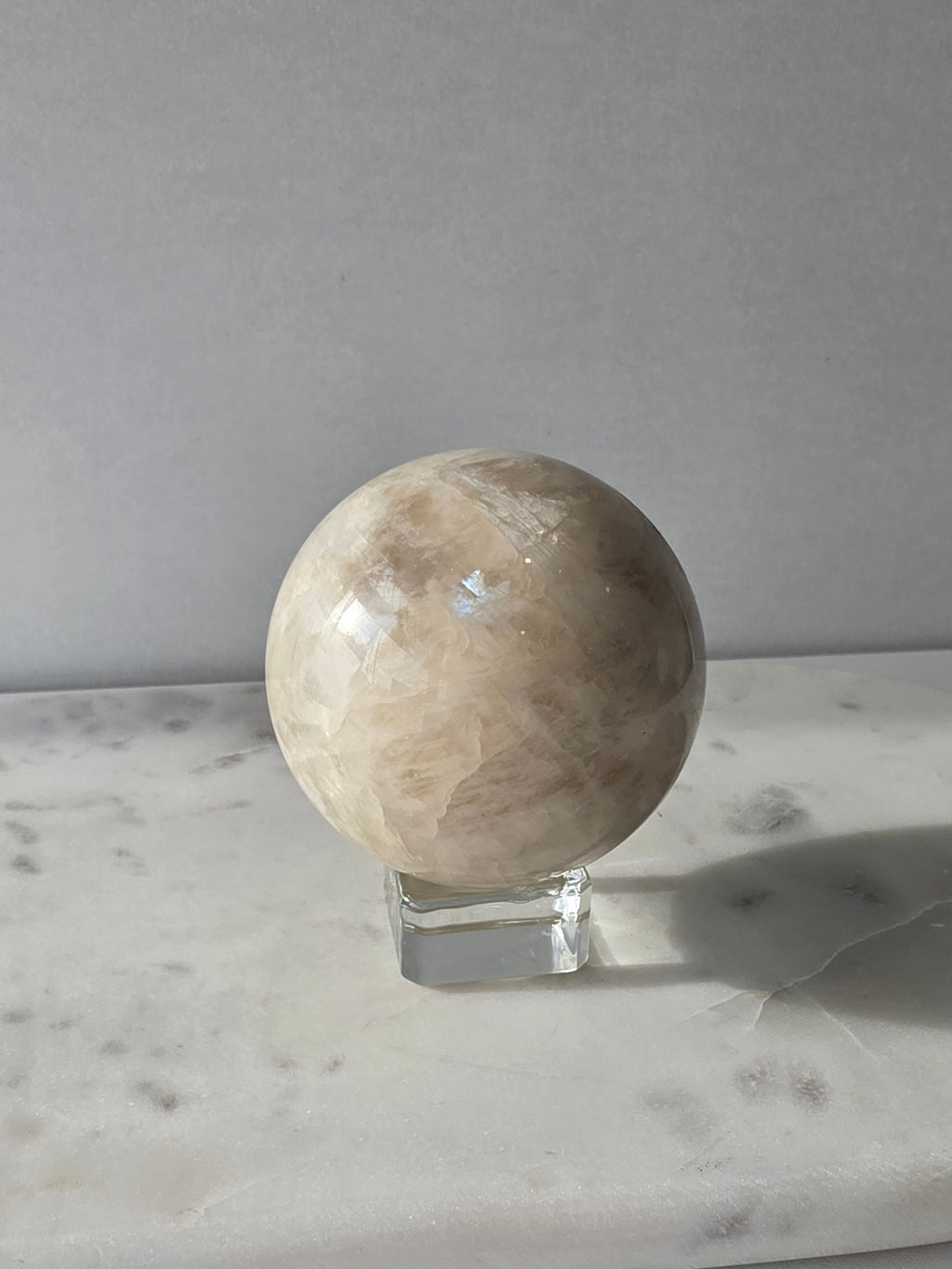 Polished Moonstone Sphere in creamy ivory, champagne and taupe-grey tones, displaying subtle natural patterns and a soft luminous sheen on a clear stand.