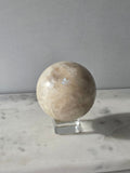 Polished Moonstone Sphere in creamy ivory, champagne and taupe-grey tones, displaying subtle natural patterns and a soft luminous sheen on a clear stand.