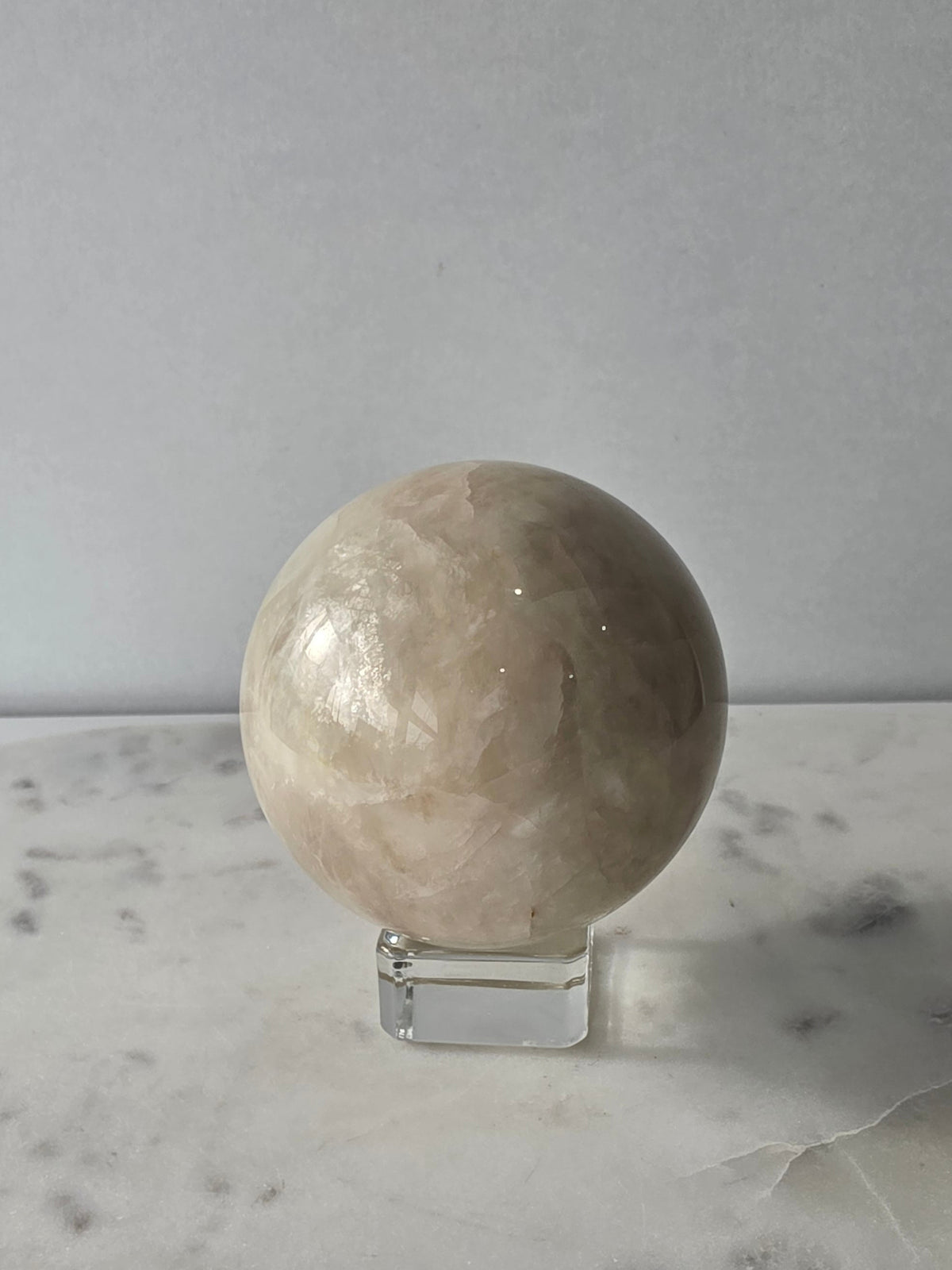 Polished Moonstone Sphere in creamy ivory, champagne and taupe-grey tones, displaying subtle natural patterns and a soft luminous sheen on a clear stand.