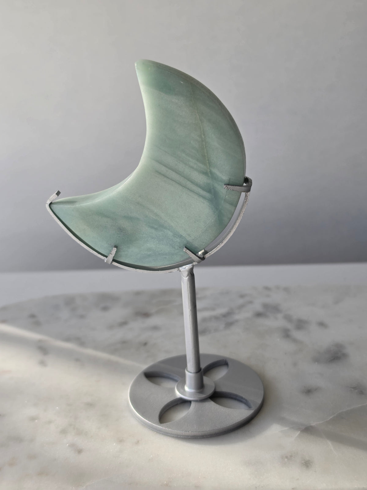 Natural Amazonite crescent moon with soft seafoam green and blue-green banding, subtle golden-brown inclusions and polished finish, displayed on a silver stand.