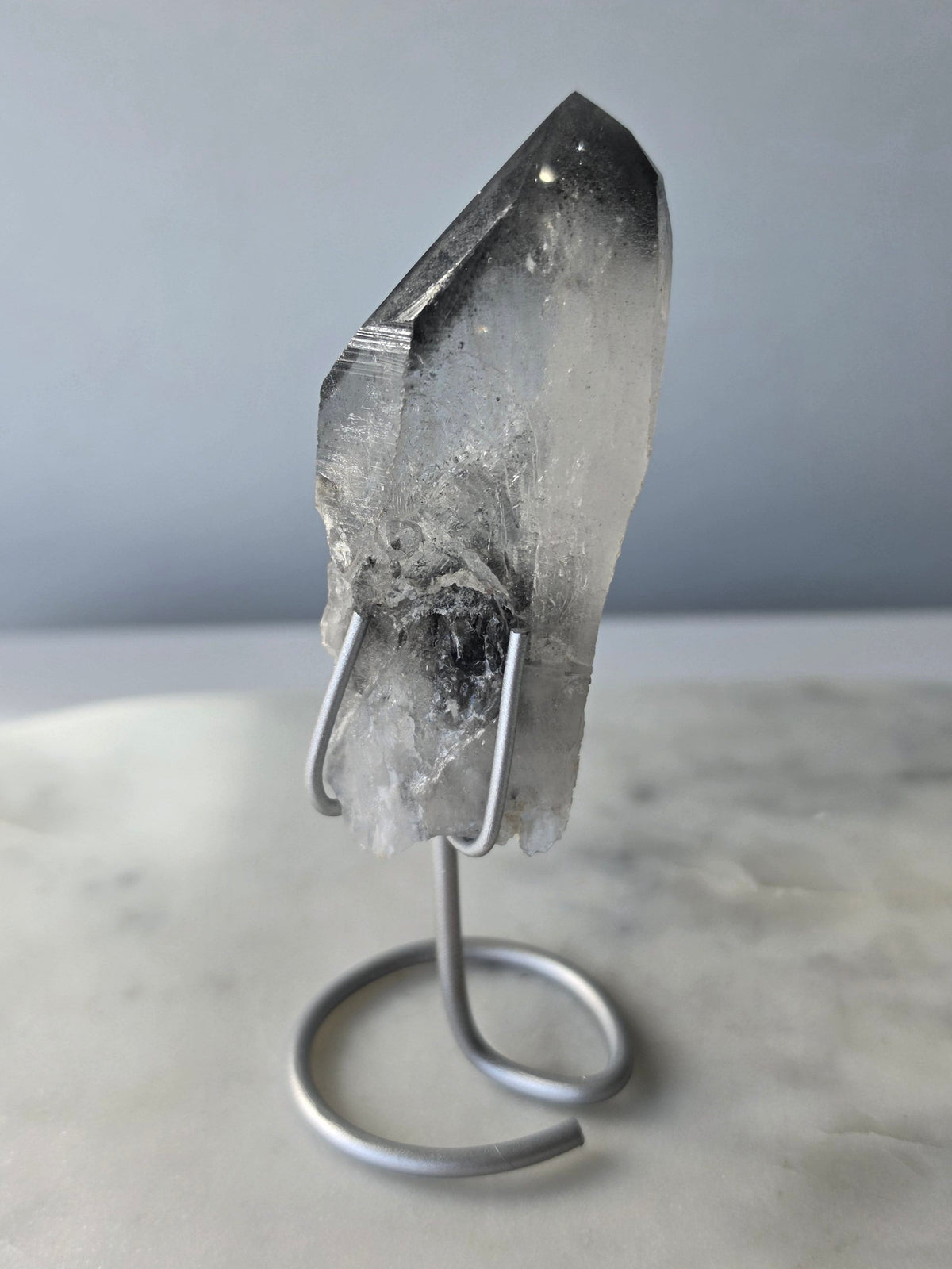 Black Phantom Lemurian Seed quartz point featuring smoky translucent crystal and dramatic charcoal-black internal phantom-like formations, displayed on a silver-toned stand.