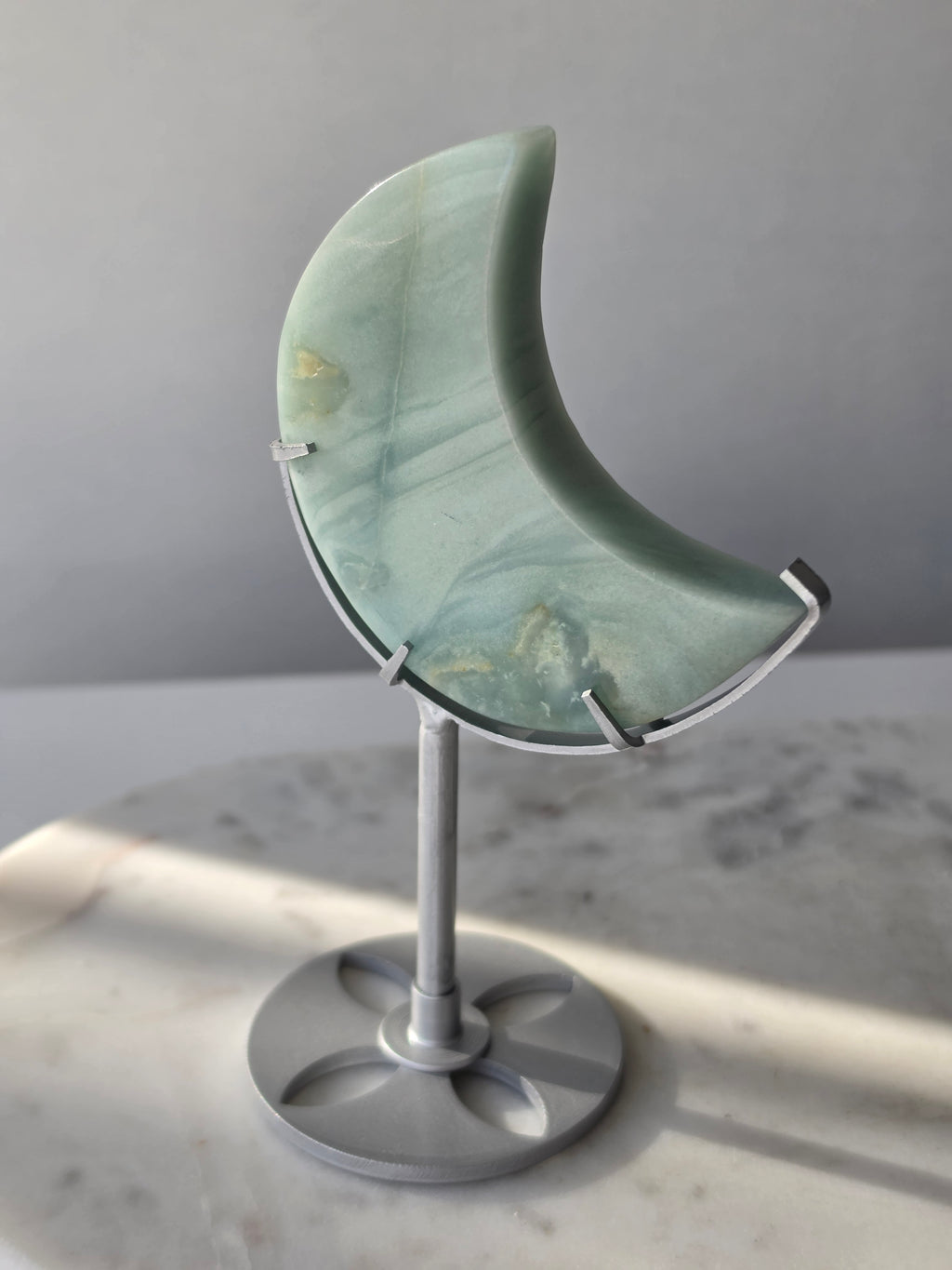 Natural Amazonite crescent moon with soft seafoam green and blue-green banding, subtle golden-brown inclusions and polished finish, displayed on a silver stand.