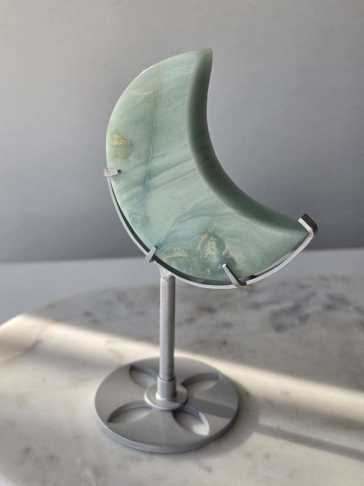 Natural Amazonite crescent moon with soft seafoam green and blue-green banding, subtle golden-brown inclusions and polished finish, displayed on a silver stand.